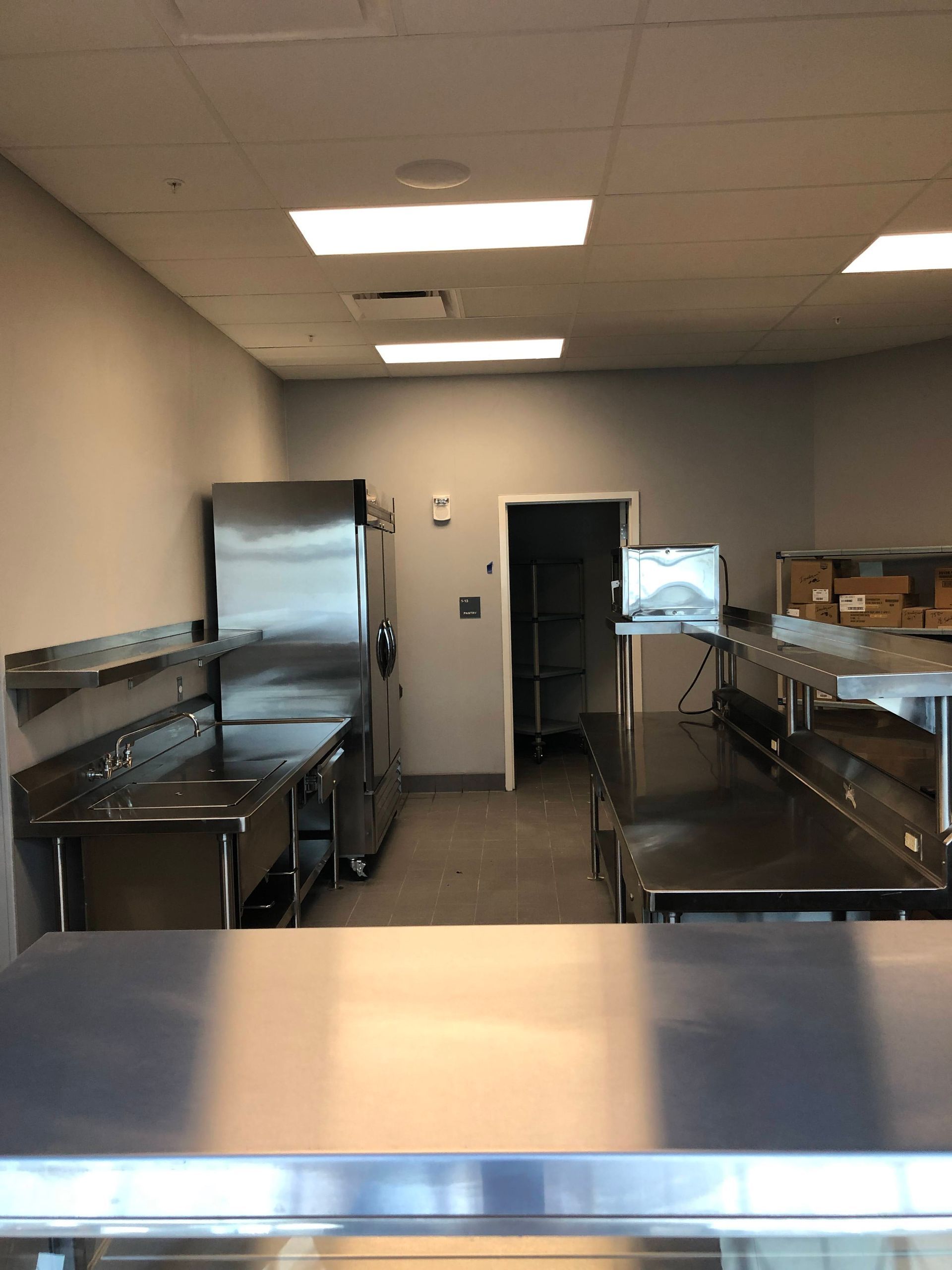 A view of a commercial kitchen featuring stainless steel prep tables, a large refrigerator, and a doorway in the back.