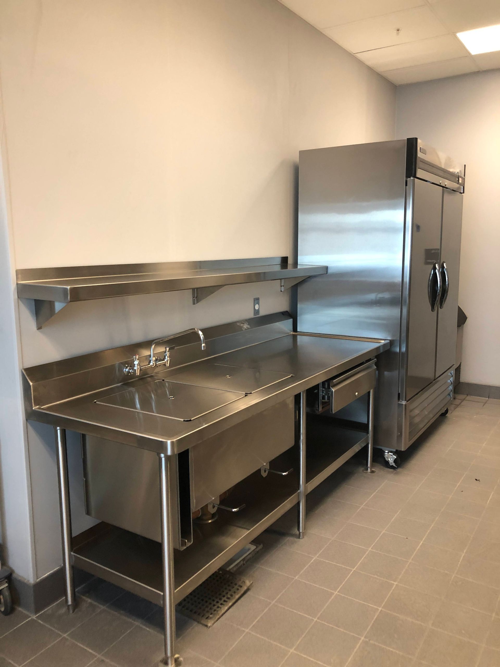 A stainless steel commercial sink with an overhead shelf and a large, upright industrial refrigerator in a kitchen space.