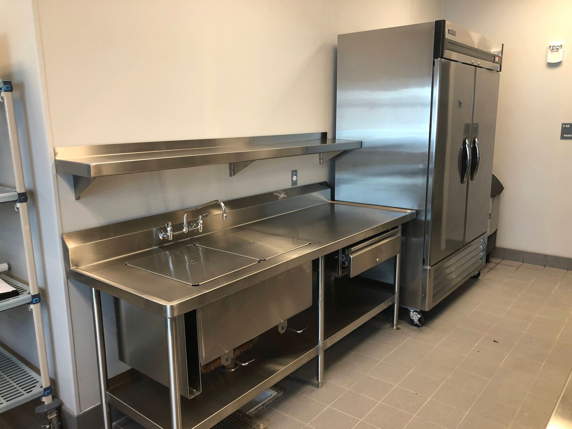 A stainless steel industrial prep table with an integrated sink and shelving next to a tall commercial refrigerator.