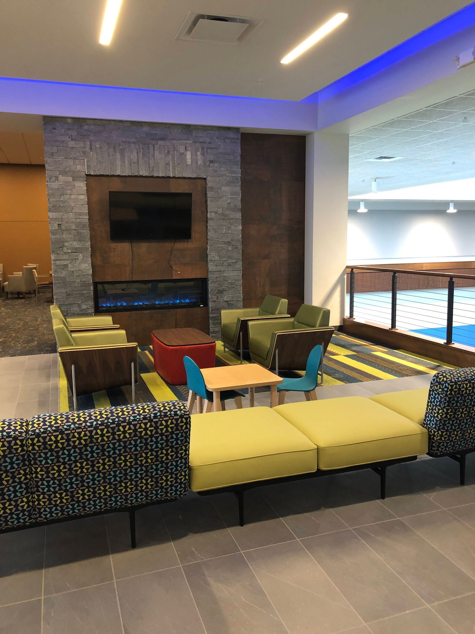 A lounge area with green armchairs, a fireplace with a mounted TV, a small table with two chairs, and a long yellow sofa.