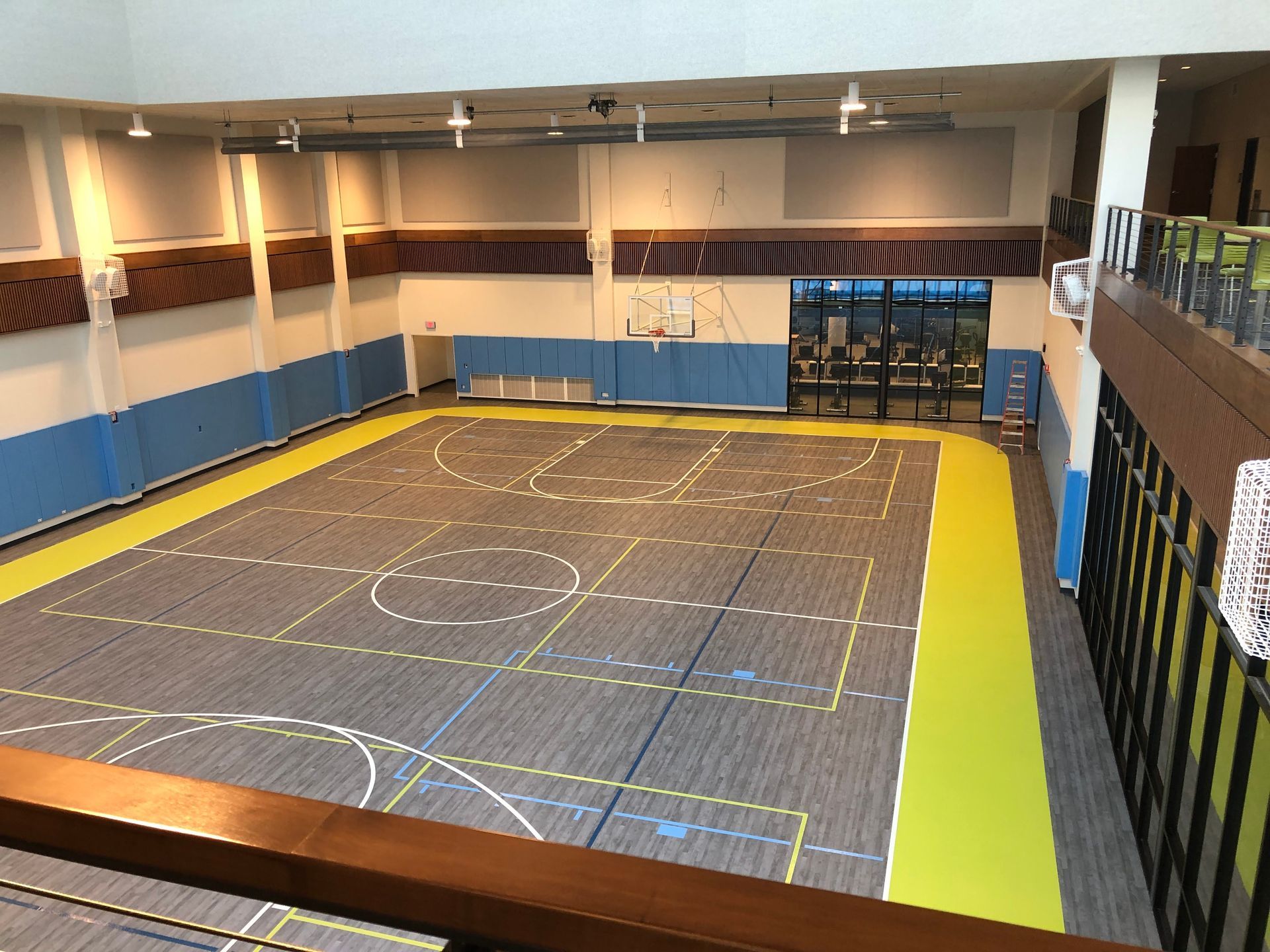 An indoor gymnasium with a gray multi-court floor, lime green borders, blue wall panels, and a mezzanine balcony.