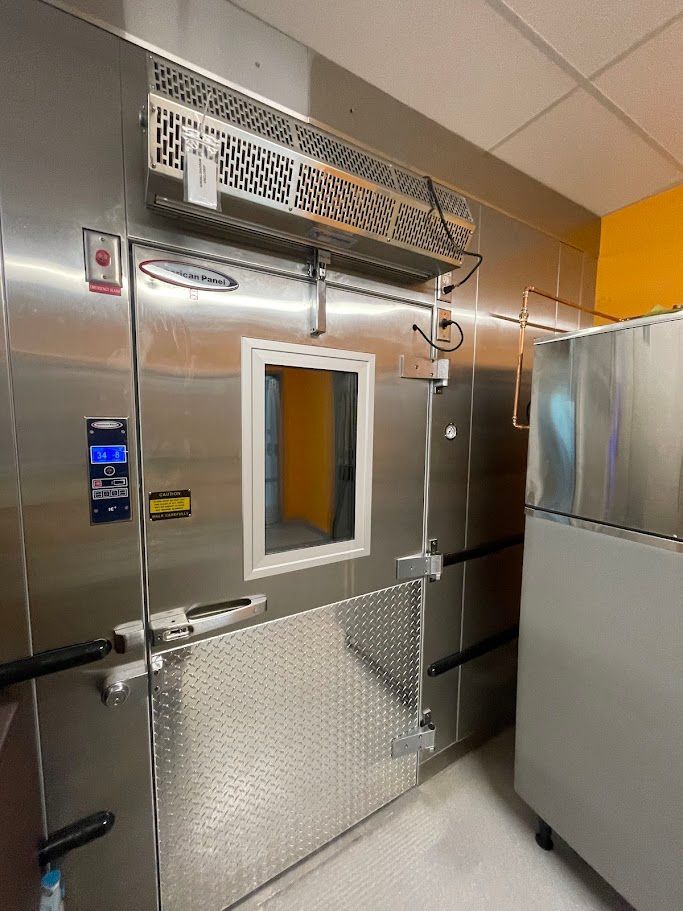 A stainless steel walk-in cooler door with a window and digital temperature control panel in a commercial kitchen.