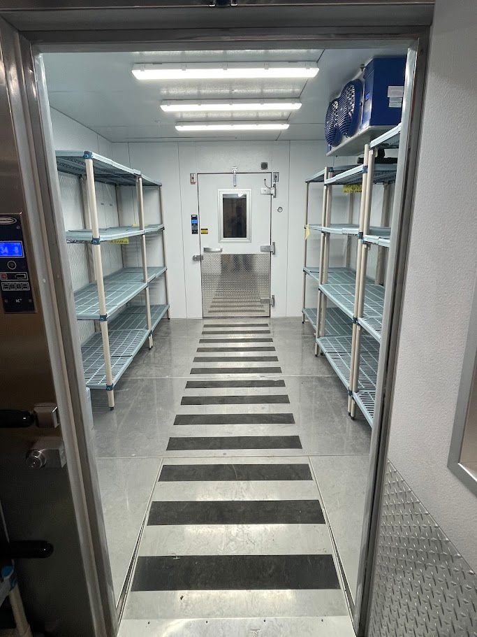 View through a doorway into a sterile, well-lit walk-in cooler with metal shelving units on both sides and a floor mat.