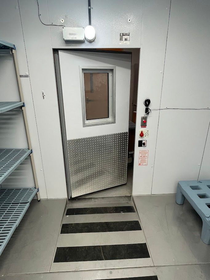 An open doorway with a metal kickplate and window, leading into a room from a storage area with shelving and floor mats.