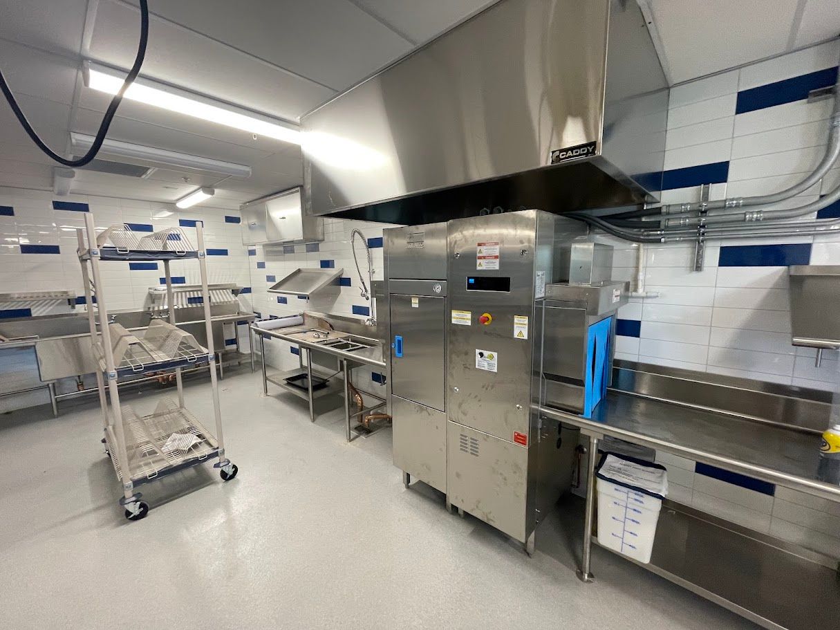 A stainless steel commercial kitchen featuring an industrial dishwasher, prep tables, and a storage rack on wheels.
