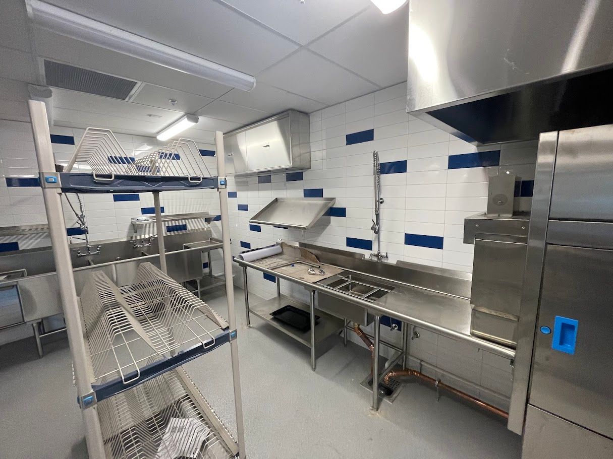 A commercial kitchen featuring stainless steel prep tables, a metal storage rack, and white tiled walls with blue accents.