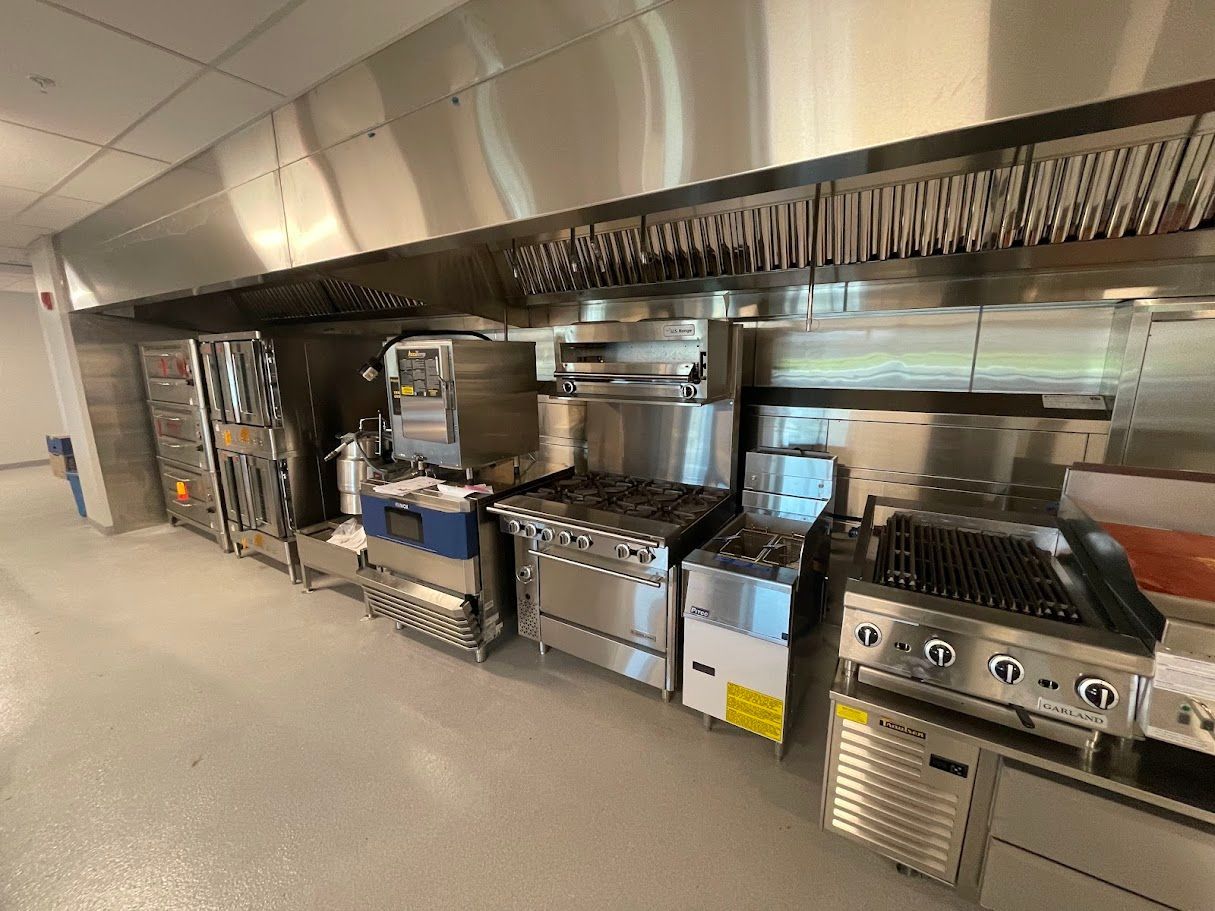 A stainless steel commercial kitchen featuring a row of stoves, ovens, and a large overhead ventilation hood.