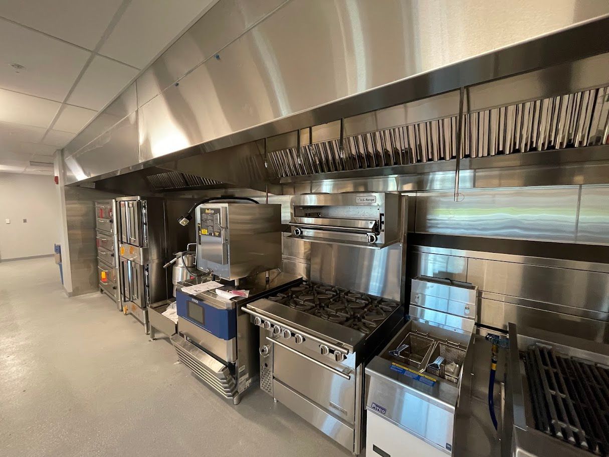 A commercial kitchen features a stainless steel vent hood over a row of industrial stoves, ovens, and grilling equipment.