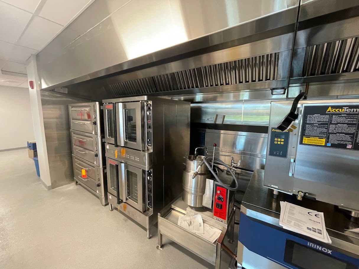 Stainless steel commercial kitchen equipment, including multiple ovens and a ventilation hood, lined up along a wall.