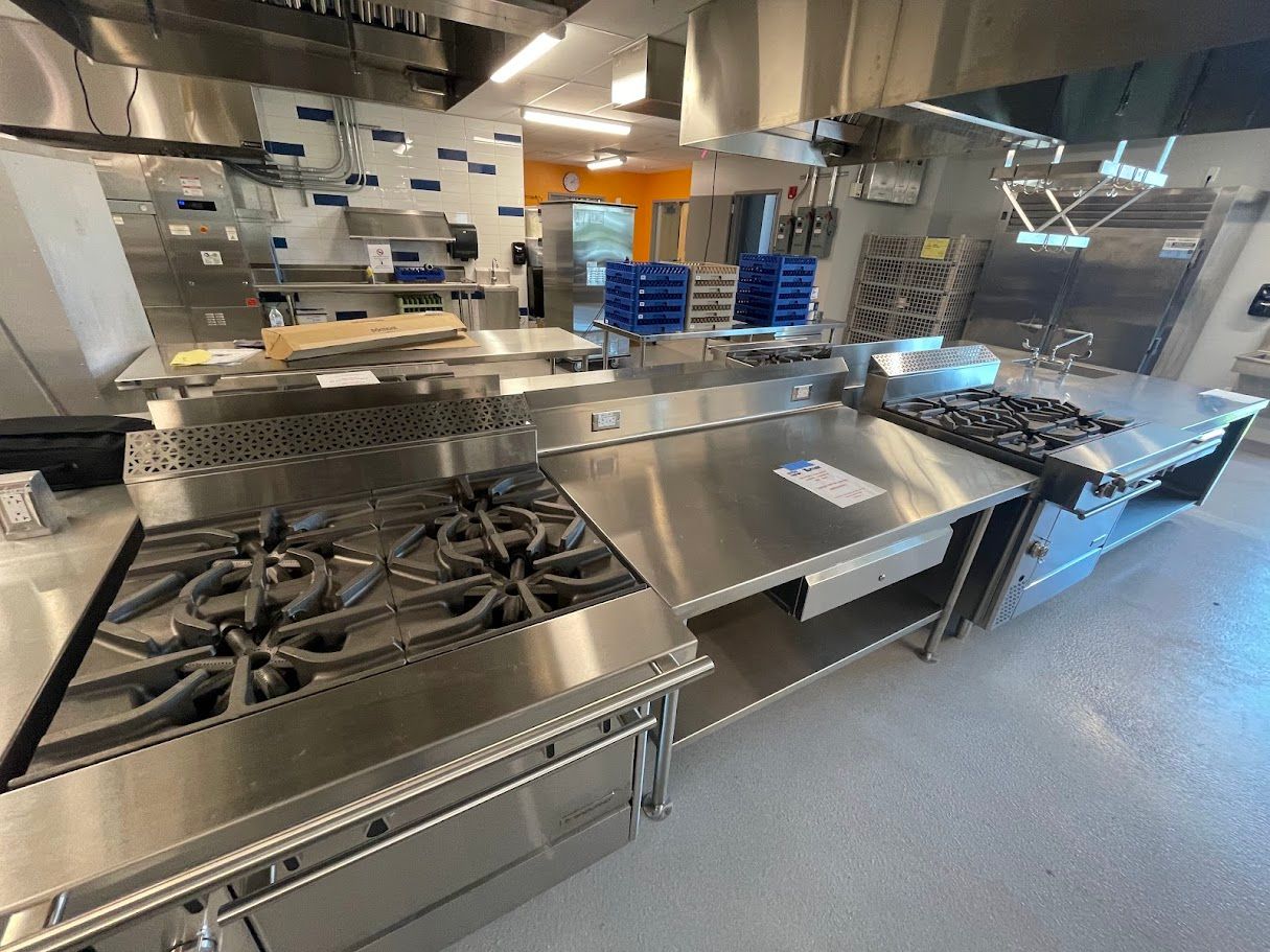 Commercial kitchen featuring stainless steel stoves, a central prep table, and work surfaces under metal exhaust hoods.