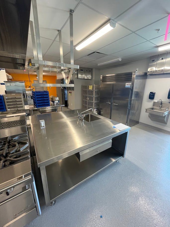 A commercial kitchen featuring a stainless steel prep table with a sink, a double-door refrigerator, and a gas stove.