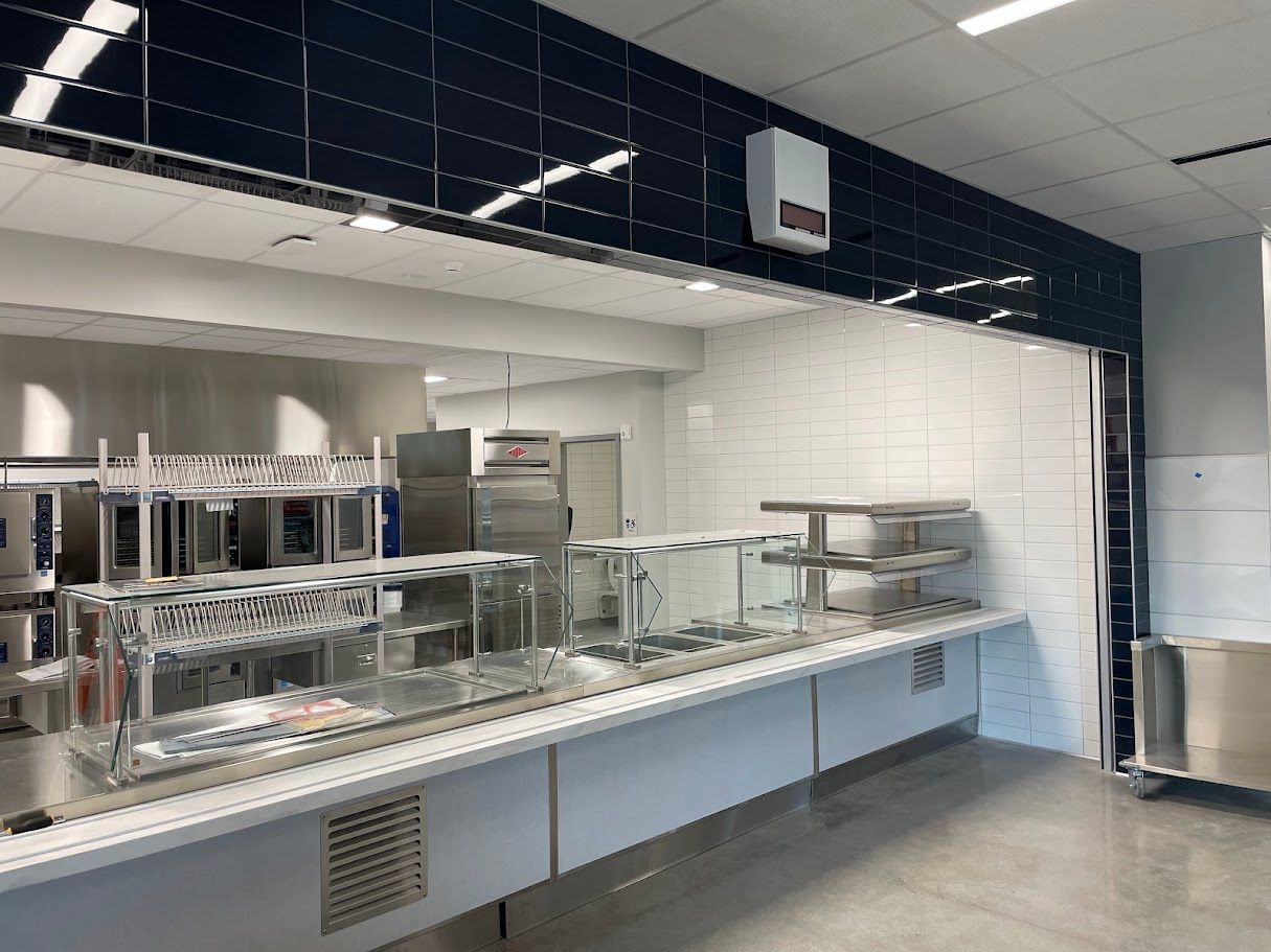 A stainless steel food serving line with shelving, sneeze guards, and heating lamps in a bright, modern commercial kitchen.