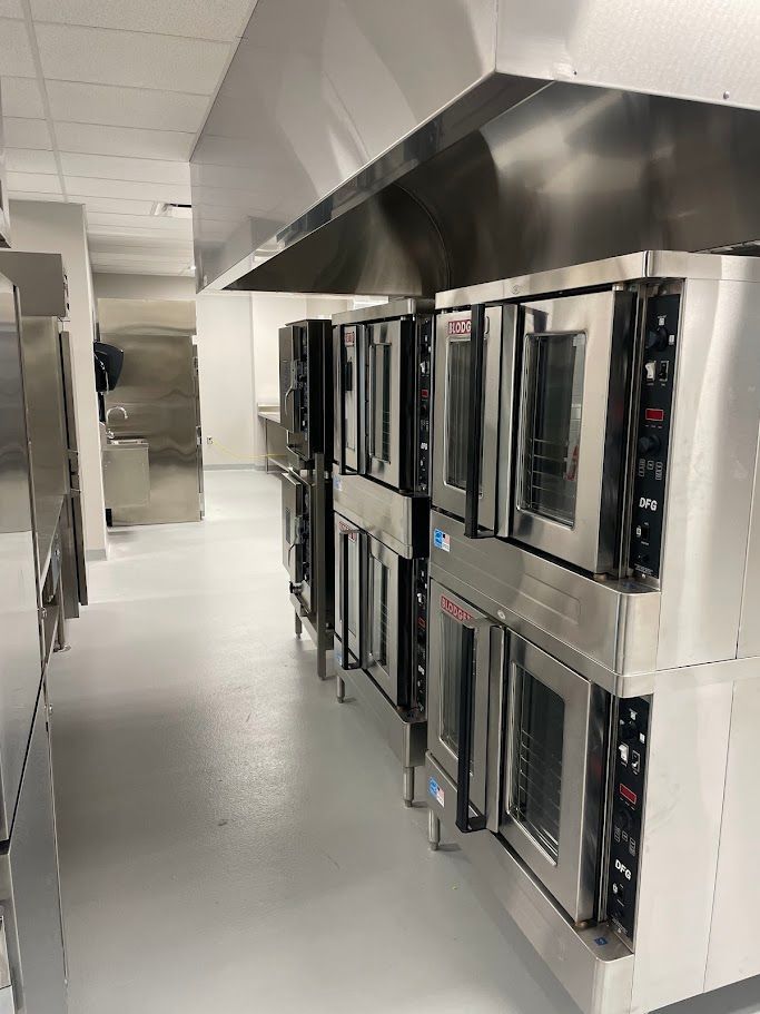 A commercial kitchen featuring rows of stainless steel industrial ovens positioned beneath a large ventilation hood.