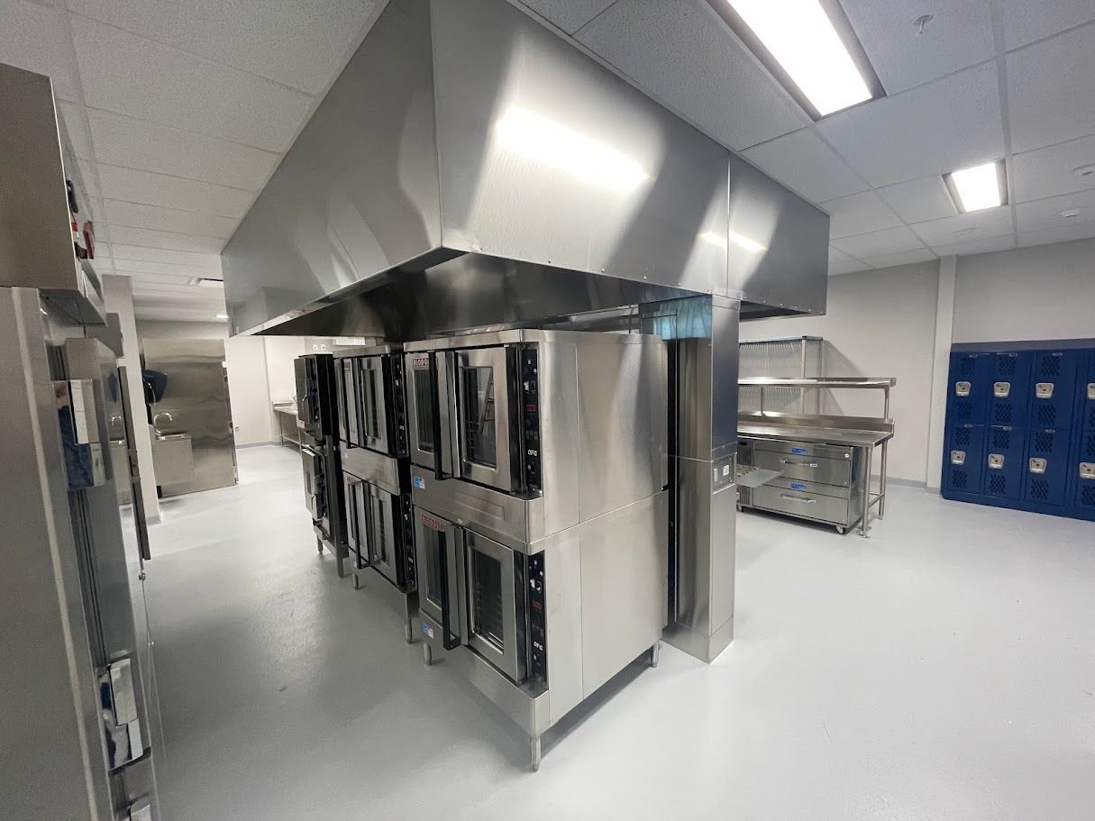 Commercial kitchen with stainless steel convection ovens under a large vent hood and a set of blue lockers.