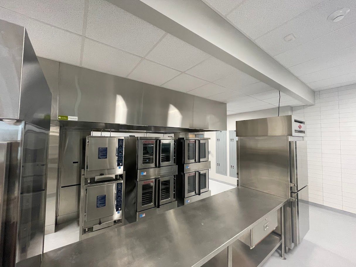 A professional stainless steel commercial kitchen featuring multiple industrial ovens, a prep table, and a refrigerator.