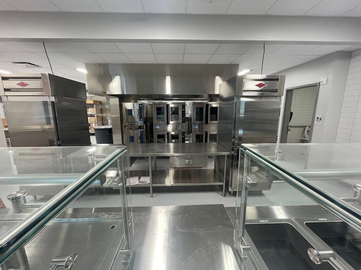 Commercial kitchen interior with stainless steel counters, industrial ovens, and a serving counter with glass guards.