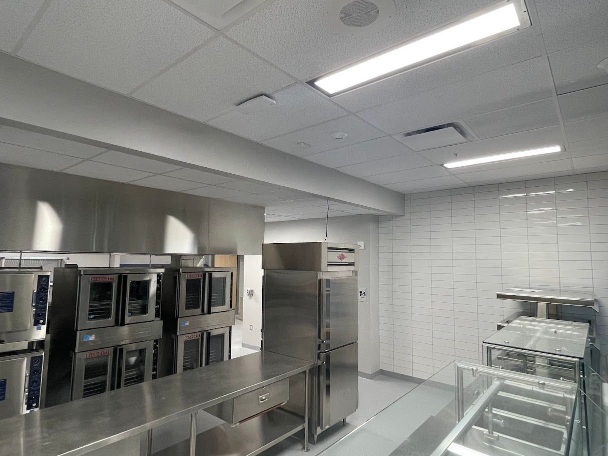 A commercial kitchen featuring stainless steel ovens, prep counters, and a white tiled wall under bright LED lighting.