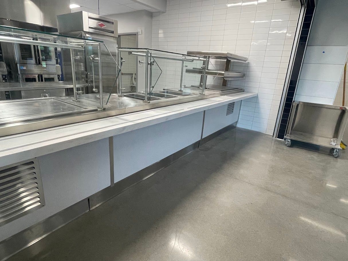A modern, stainless steel food service counter and sneeze guard in a brightly lit kitchen with a white brick wall.