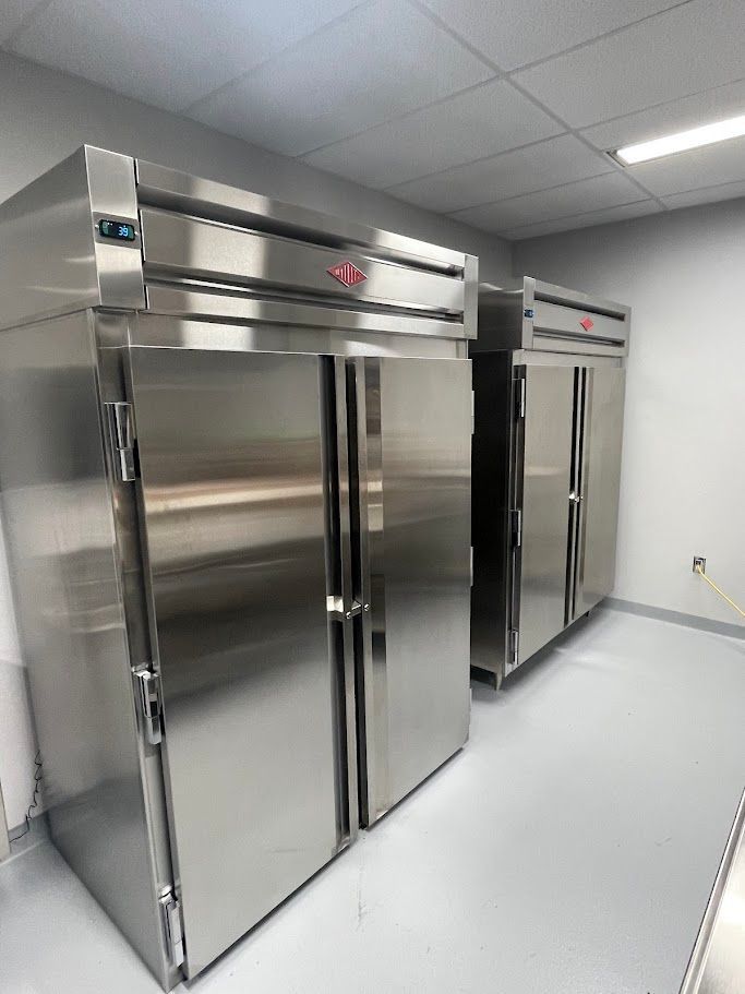 Two stainless steel commercial refrigerators standing side-by-side in a room with light grey walls and flooring.