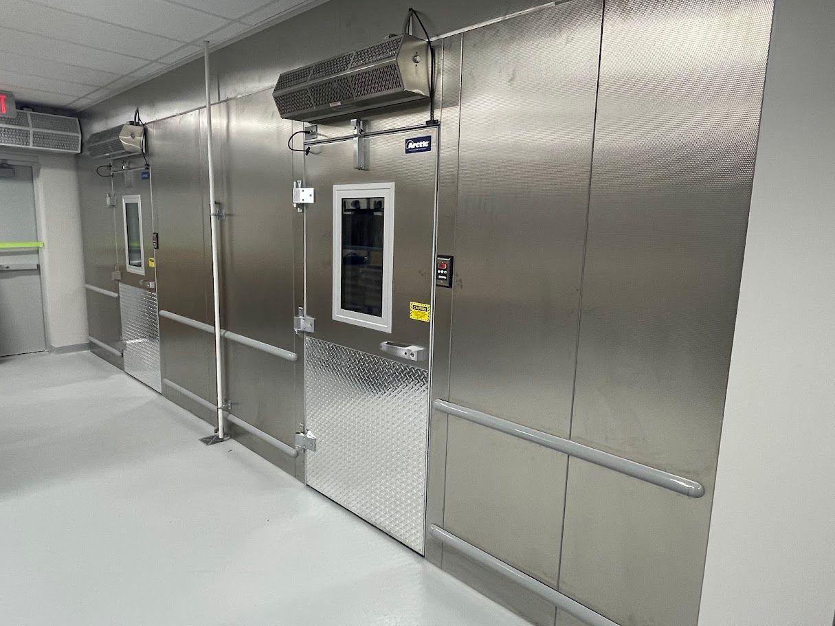 Two stainless steel industrial walk-in freezer doors with diamond-plated kickplates in a clean, brightly lit facility.