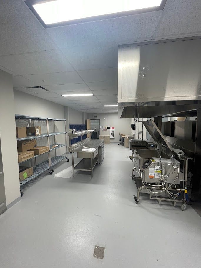 A commercial kitchen featuring metal shelving with boxes, stainless steel prep tables, and cooking equipment under a hood.