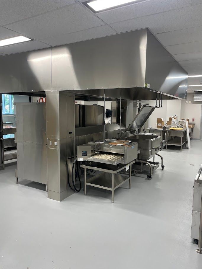 A stainless steel commercial kitchen hood hangs over equipment, including a countertop griddle and prep tables in a room.