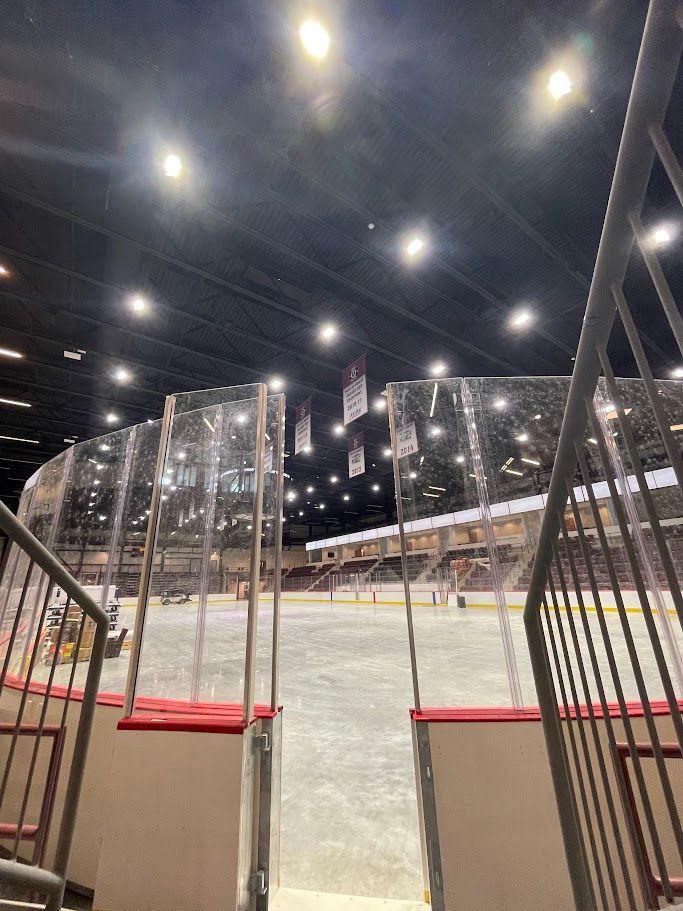 A view from the stands overlooking an empty indoor ice hockey rink with glass dasher boards and overhead arena lighting.