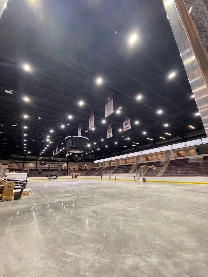 Empty indoor ice hockey arena with dark ceilings, stadium seating, and overhead lights.