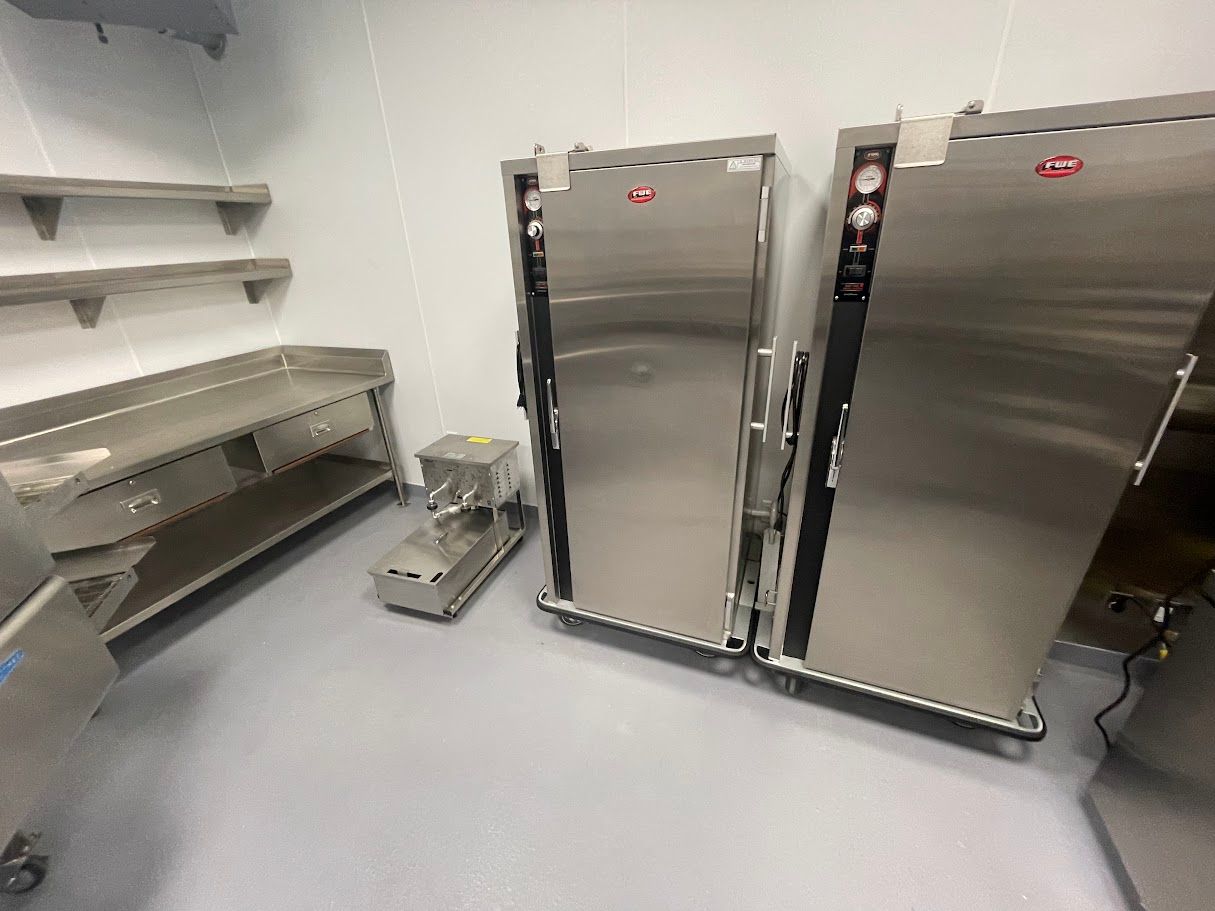 Two stainless steel industrial ovens stand next to a prep table and a small food processor in a sterile commercial kitchen.