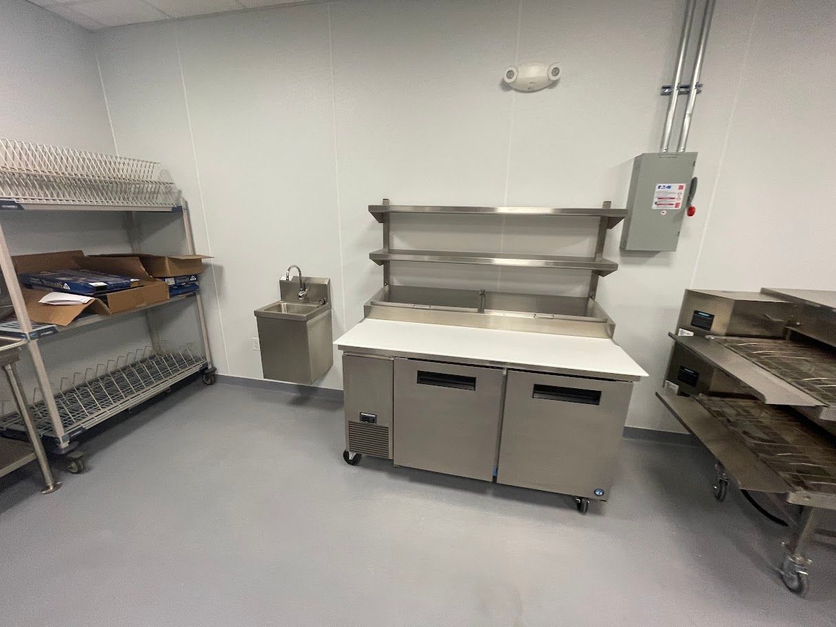 A sterile commercial kitchen room featuring metal shelving, a small sink, a prep table, and a stainless steel counter.