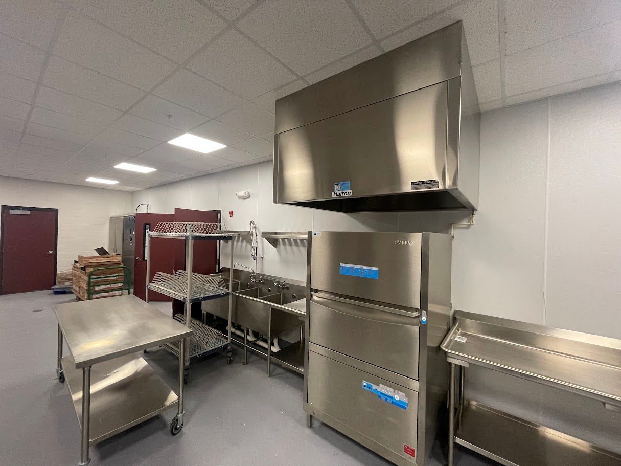 A commercial kitchen with stainless steel prep tables, a sink, and a large dishwashing machine under a ventilation hood.