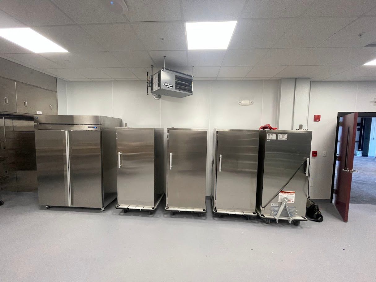 A row of stainless steel commercial refrigerators and warming cabinets in a brightly lit, industrial kitchen space.