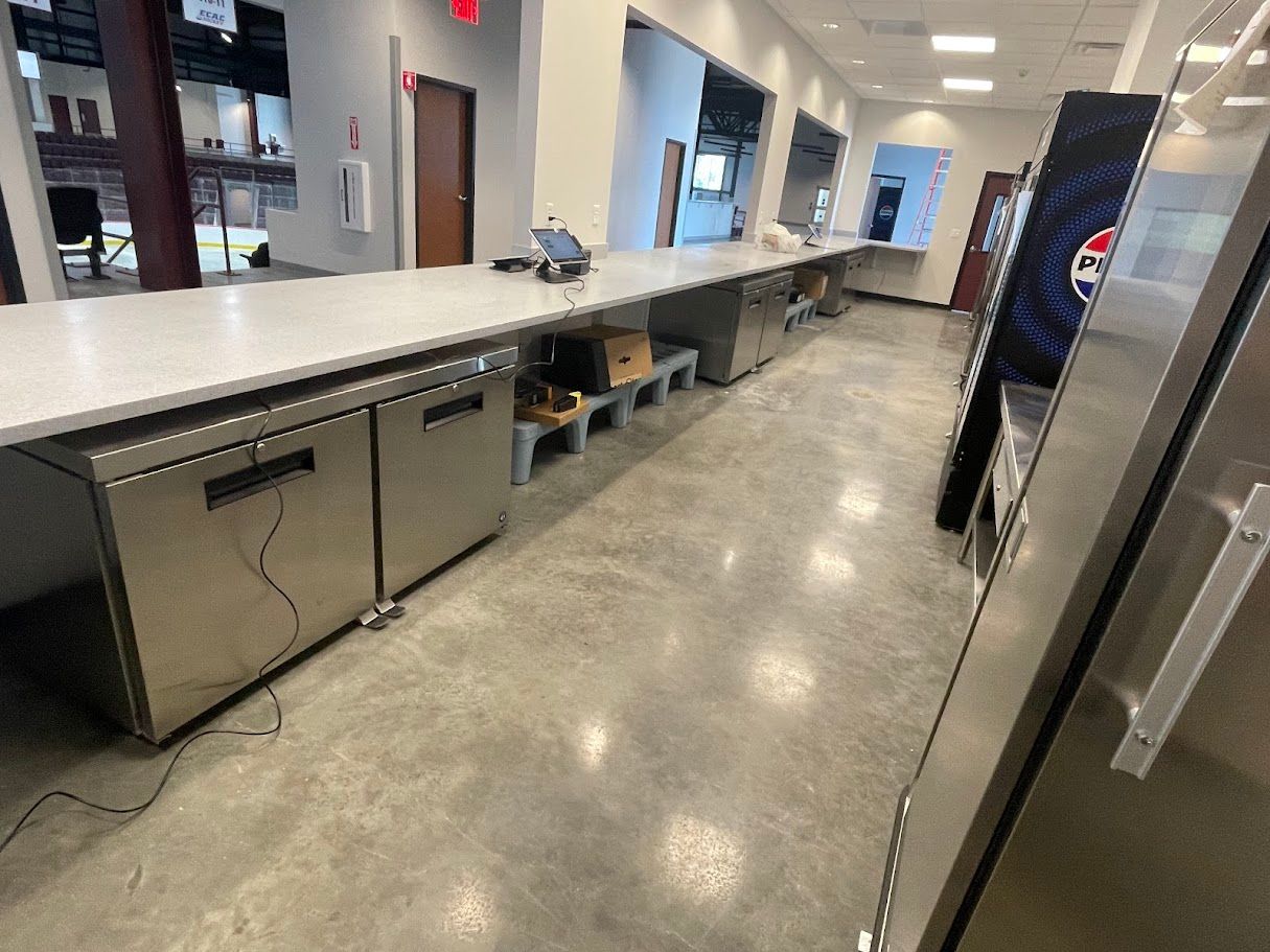 A long stainless steel counter with under-counter refrigerators, set against a concrete floor in a commercial kitchen space.
