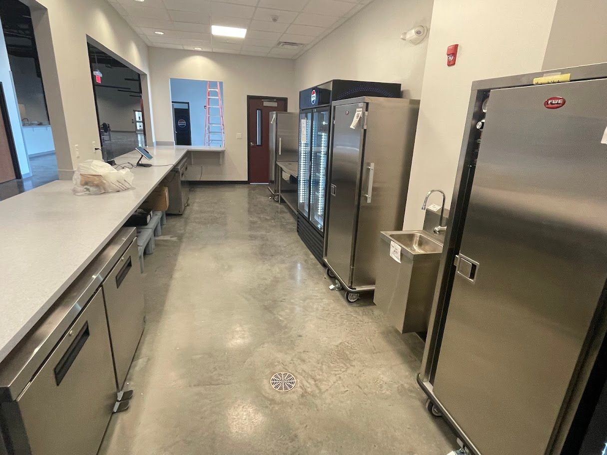 A modern, industrial-style kitchen featuring stainless steel prep tables, a walk-in cooler, and polished concrete floors.