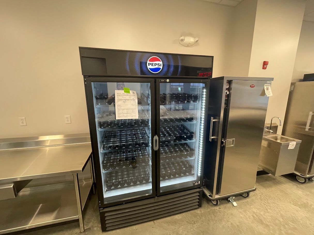 A double-door Pepsi glass-front refrigerator stands in a commercial kitchen next to metal cabinets and a prep table.