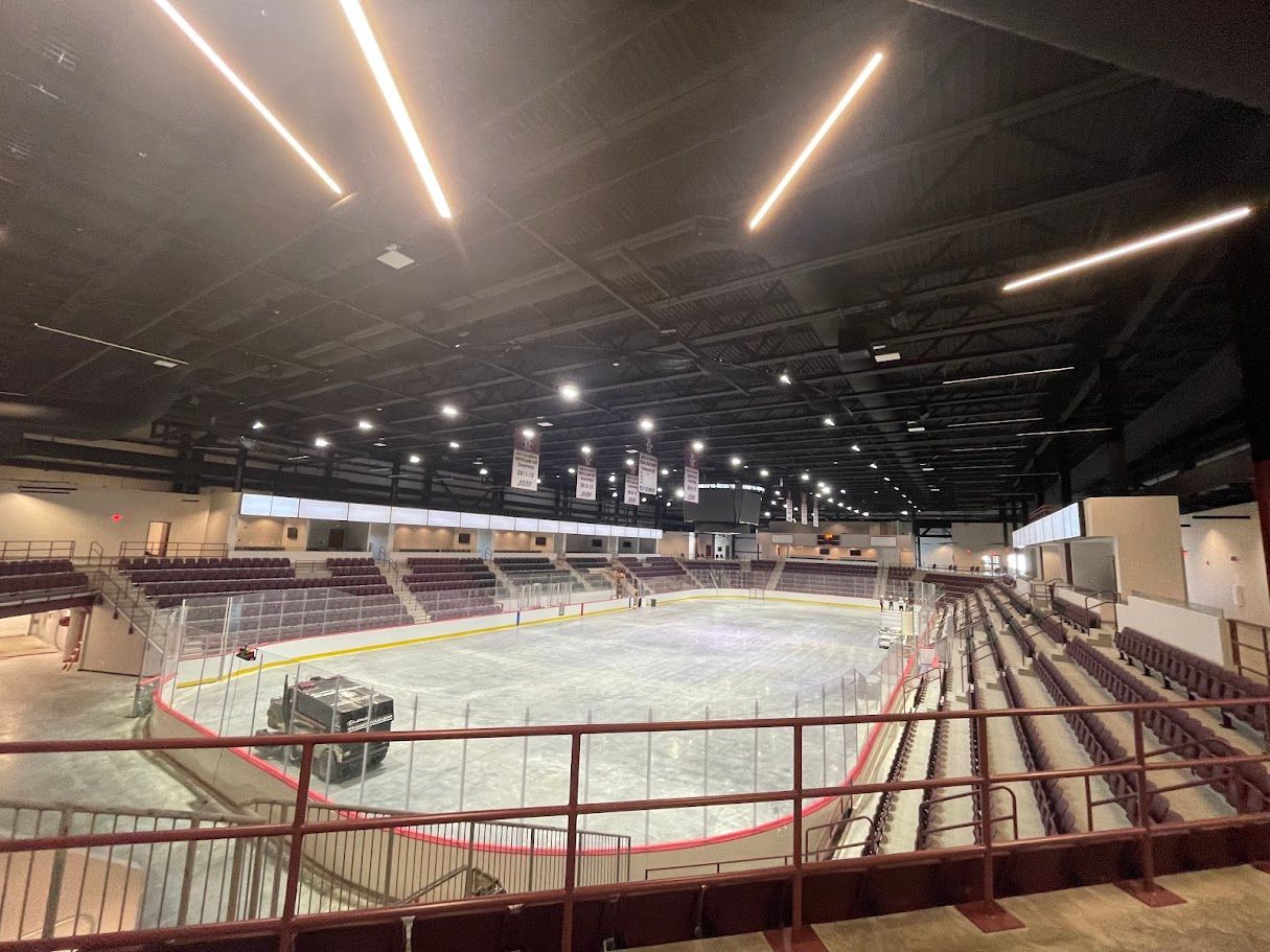 A wide-angle view of an indoor ice hockey rink with stadium seating, a resurfacing machine on the ice, and overhead lighting.