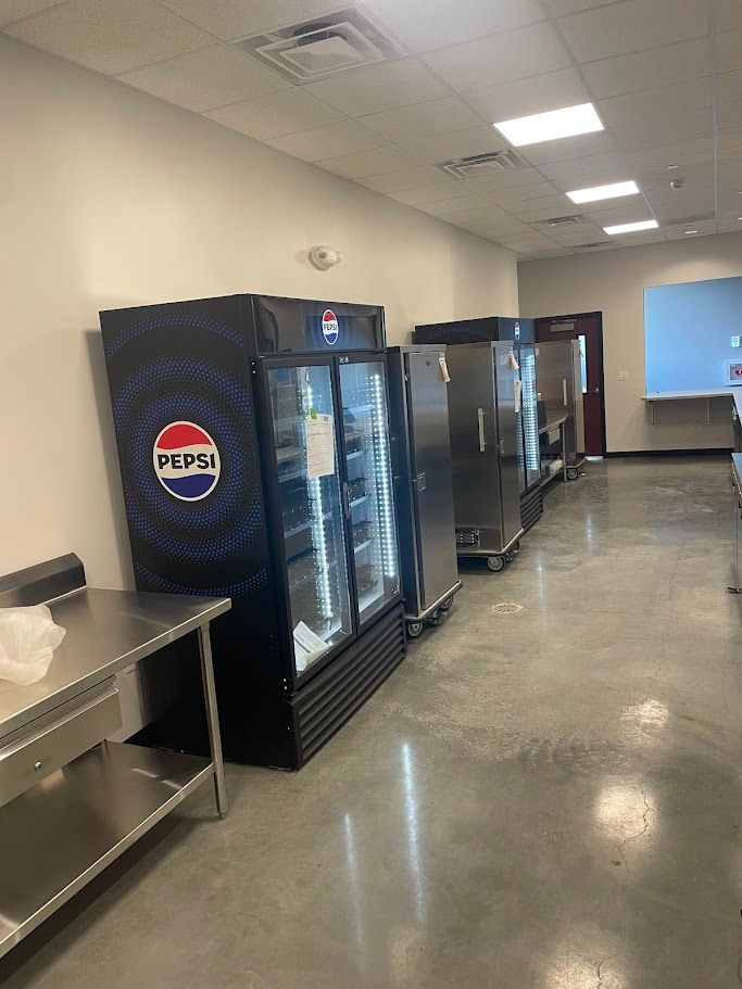 A Pepsi-branded glass-door refrigerator stands next to stainless steel prep equipment in a commercial kitchen.