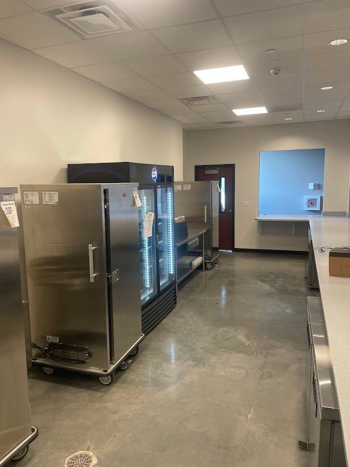 A commercial kitchen featuring stainless steel refrigerators, a service counter, and a door on a grey concrete floor.