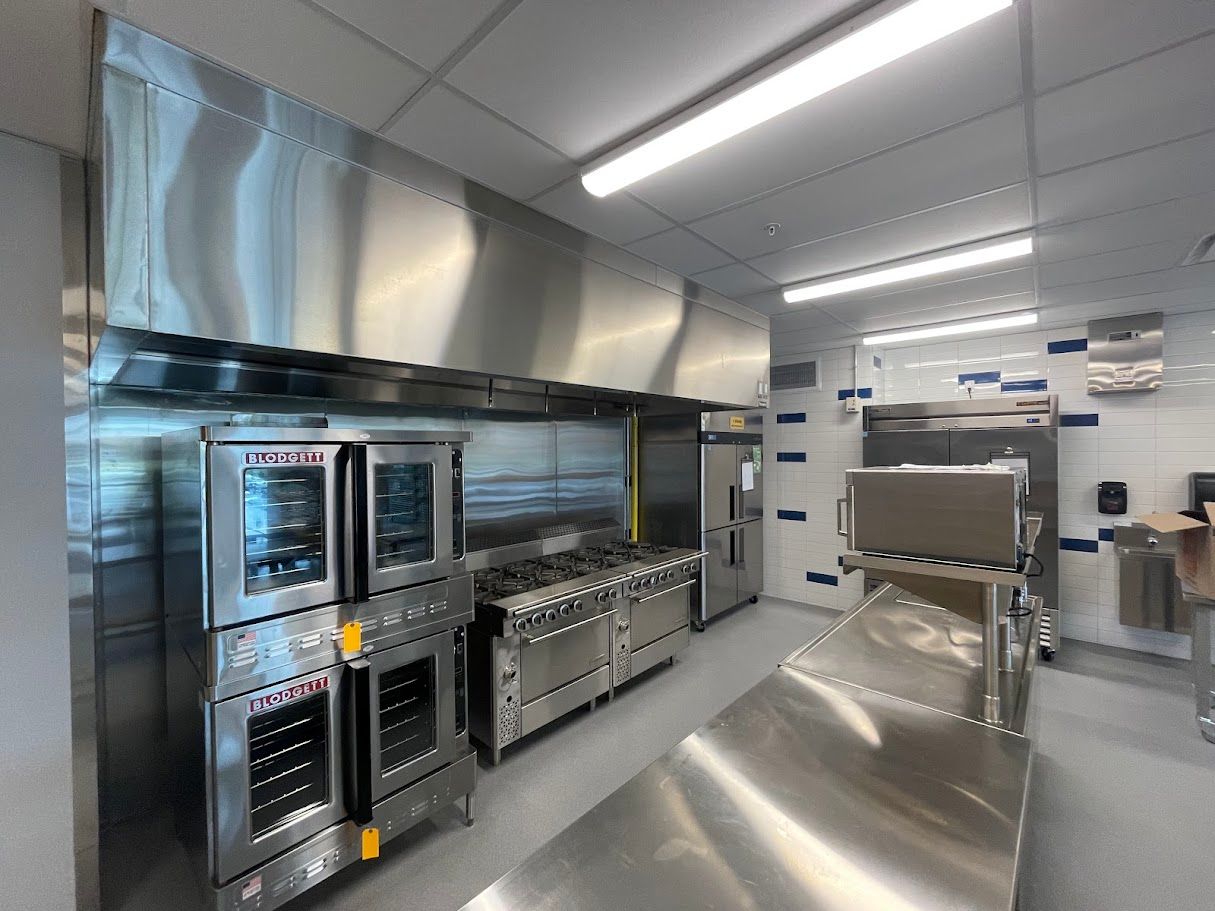 A bright, modern commercial kitchen featuring stainless steel appliances, a large exhaust hood, and a metal prep table.