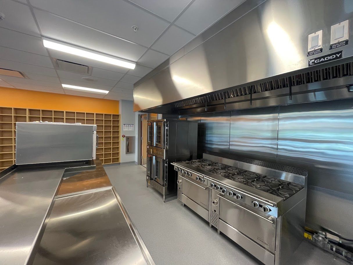 A professional commercial kitchen featuring stainless steel stoves, a ventilation hood, and shelving against an orange wall.