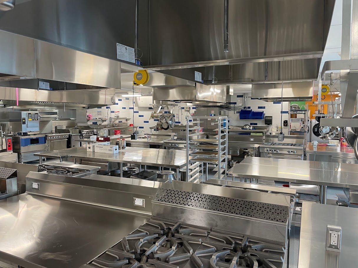 A spacious, professional industrial kitchen with stainless steel countertops, multiple stoves, and overhead vent hoods.