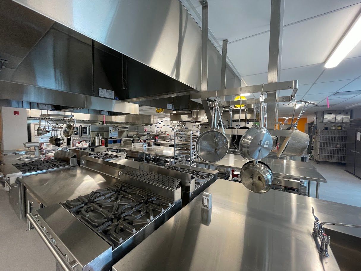 A wide, brightly lit professional kitchen with stainless steel countertops, industrial stoves, and hanging cookware.