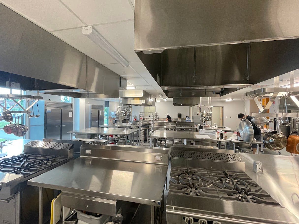A stainless steel commercial kitchen featuring multiple stoves and ventilation hoods, with people working in the background.