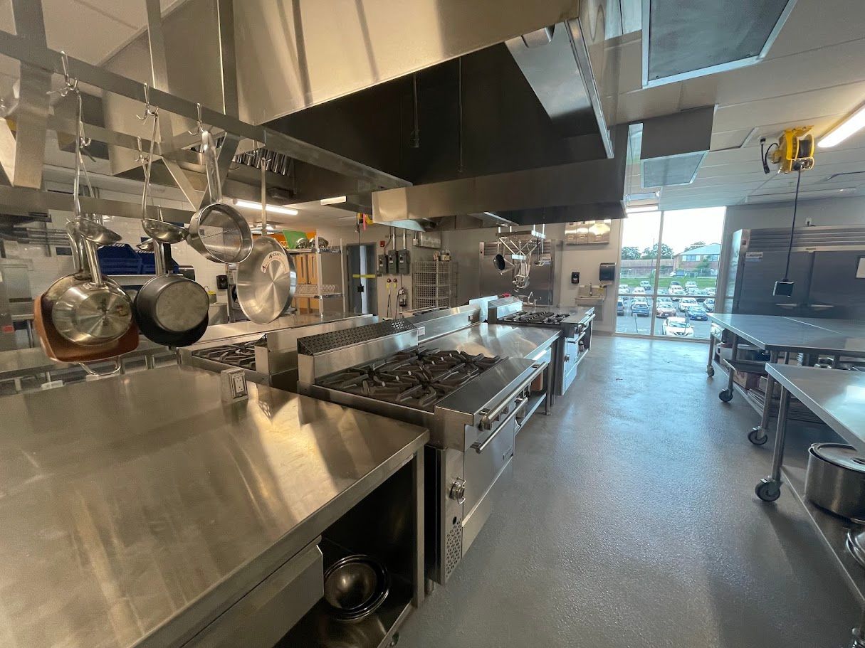 A wide-angle view of a commercial kitchen featuring stainless steel stoves, prep tables, and pots hanging overhead.