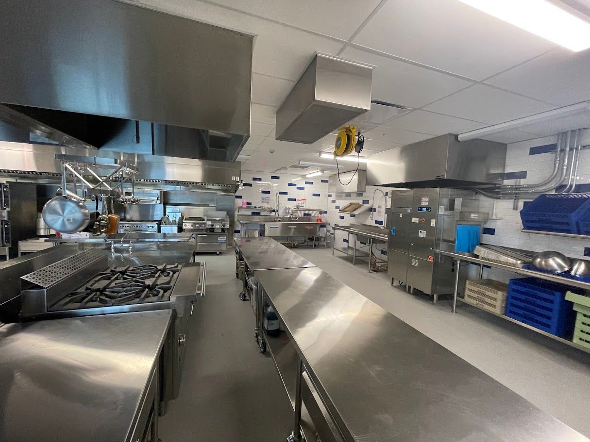 A bright, industrial commercial kitchen with stainless steel countertops, a large stove, and metal ventilation hoods.