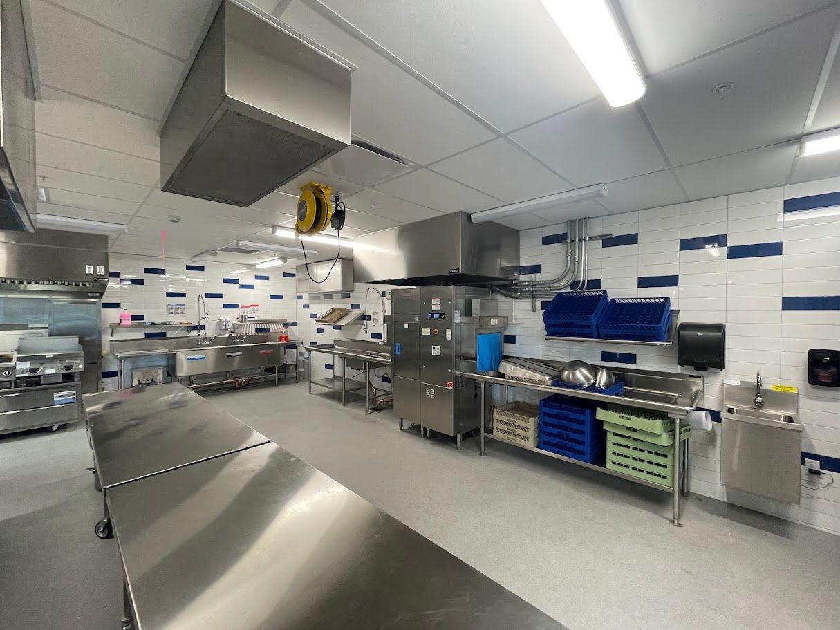 A commercial kitchen featuring stainless steel work tables, large food storage refrigerators, and shelves with blue bins.