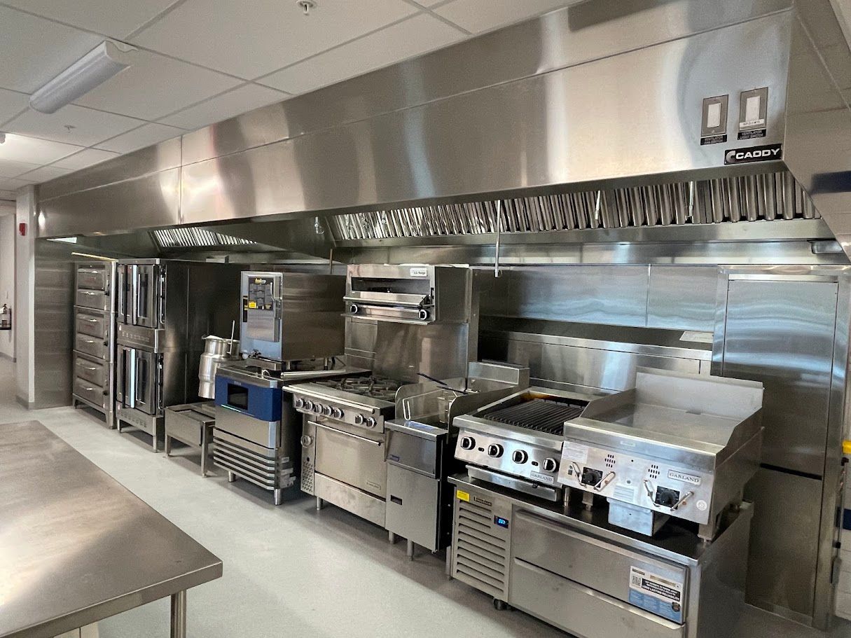 A stainless-steel commercial kitchen lineup featuring an overhead vent hood, stove ranges, grills, and ovens.