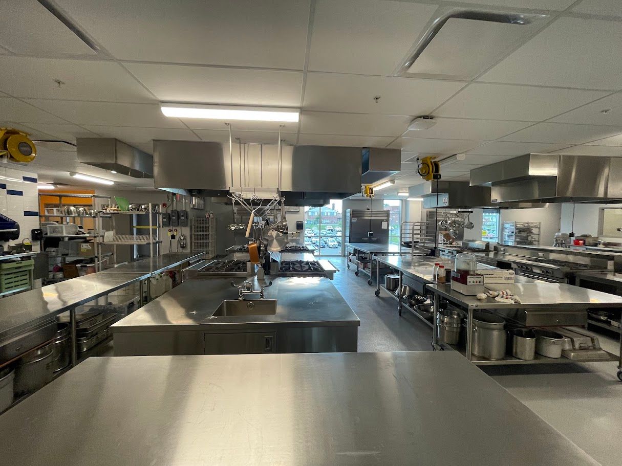A spacious, stainless steel commercial kitchen featuring multiple prep stations, workstations, and overhead vent hoods.