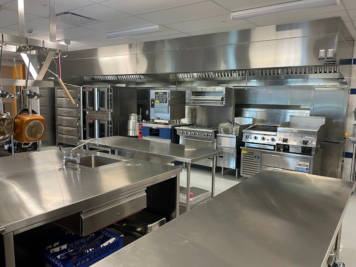 A commercial kitchen featuring stainless steel work tables, a large overhead hood, and various cooking appliances.