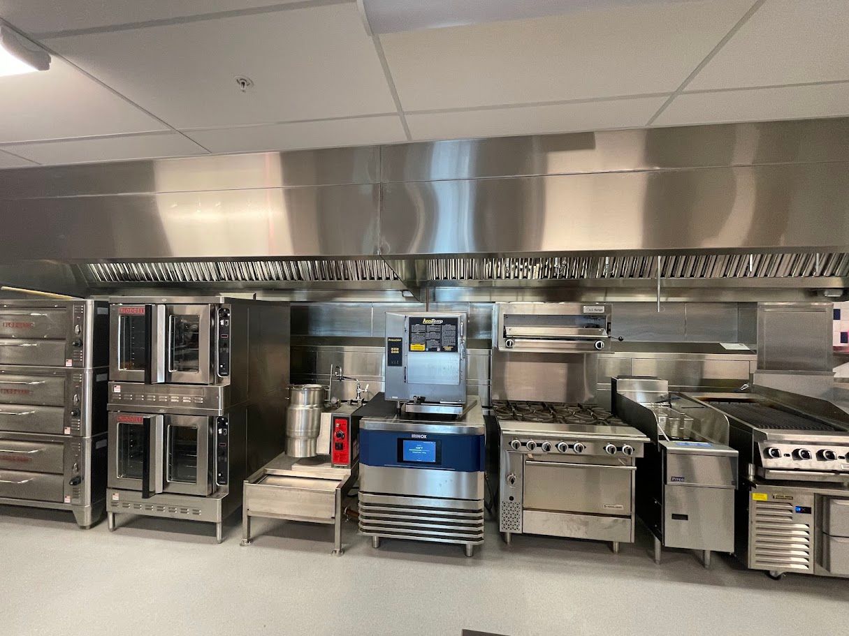 A row of industrial stainless steel kitchen equipment, including ovens, a range, and a grill, beneath a metal exhaust hood.